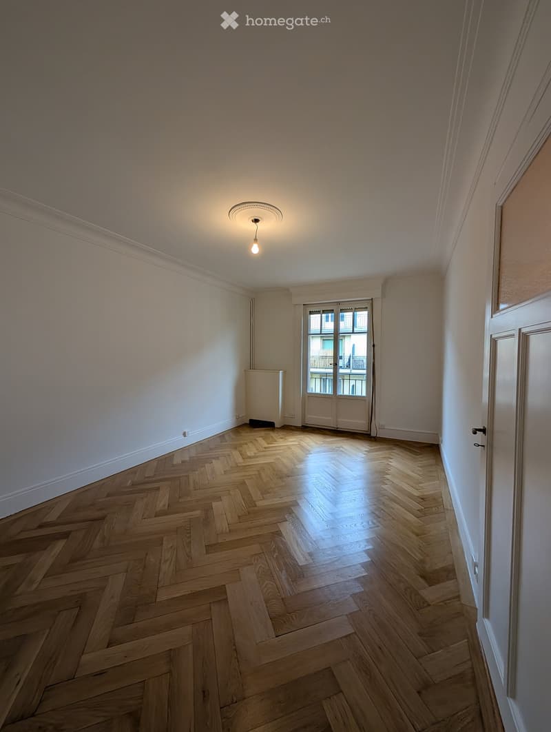 Empty room with wooden herringbone flooring, white walls, and a ceiling light fixture. A door leads to a balcony with a view of another building.
