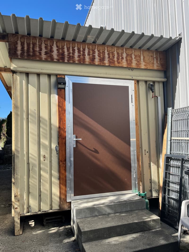 Exterior view of a small, single-story building with a metal door, wooden trim, and a concrete staircase leading up to it. The building has a weathered appearance with visible wear and tear on the siding.