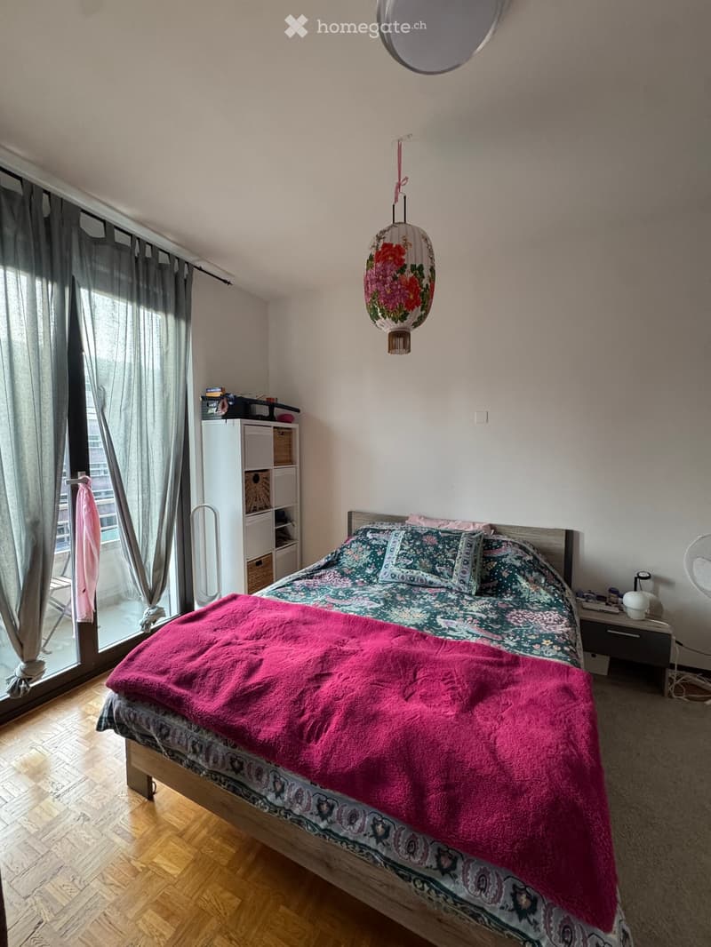 Cozy bedroom with a wooden bed, colorful bedding, pink throw, nightstand, mirror, and a hanging lantern. Large window with curtains and a view of the outside.