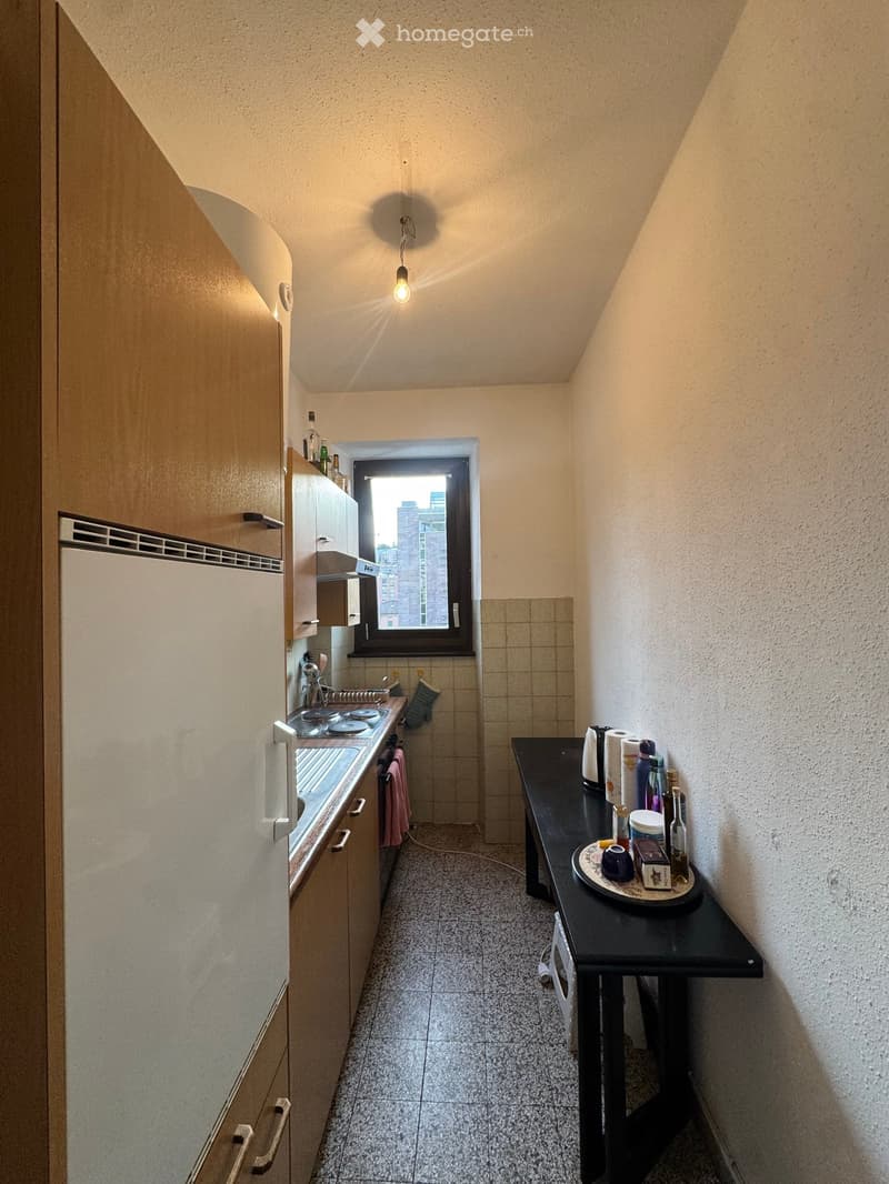 Compact kitchen with light-colored cabinets, a sink, stove, and a small table with various items on it. The space features tiled flooring and a window offering a view of the city.