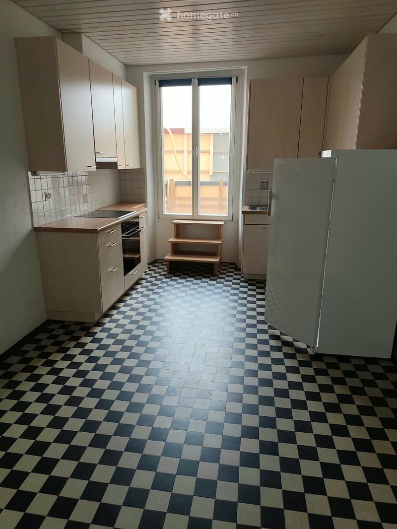 Kitchen with checkered floor, cabinets, refrigerator, oven, and a small wooden stool near the window.