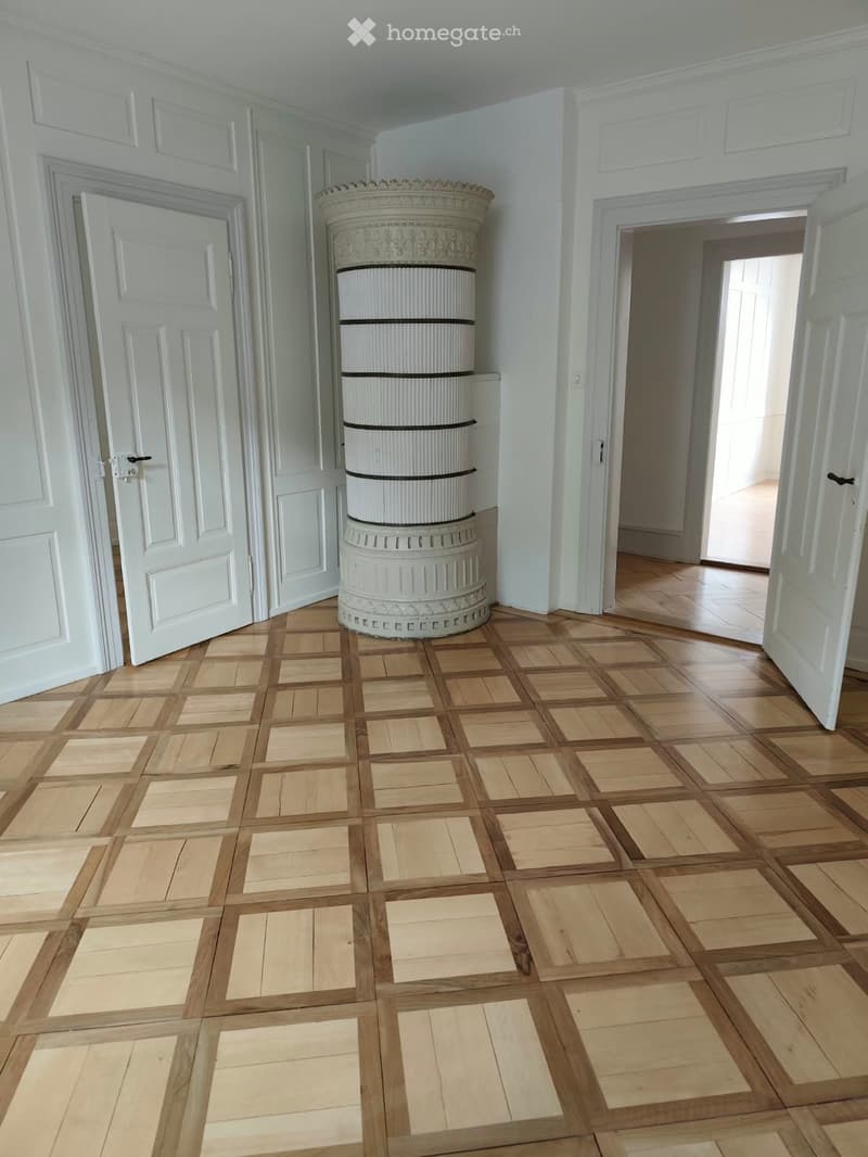 Empty room with wooden parquet flooring, white walls, and a decorative column in the center. Two open doors lead to adjacent spaces.