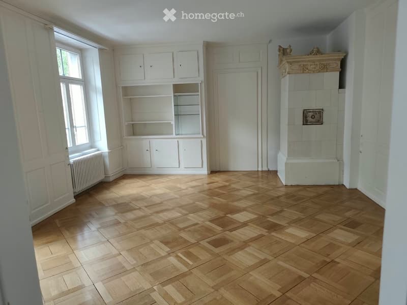 Empty room with wooden parquet flooring, white walls, built-in shelving unit, fireplace, and large windows allowing natural light.