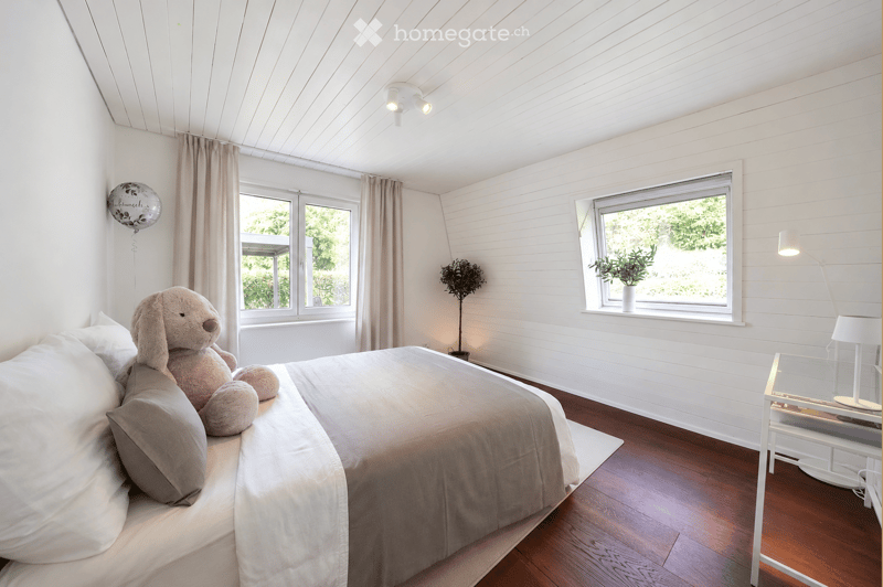 Bright bedroom with white walls, wooden floor, large windows with curtains, bed with pillows and a large stuffed animal, nightstand with lamp, small plant on windowsill, and a balloon on the wall.