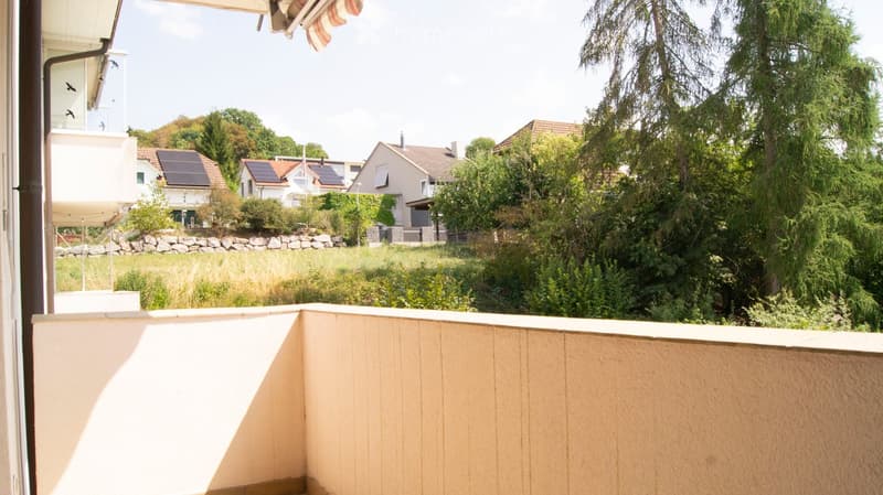 Balcony with wooden railing, overlooking a green landscape with houses and trees in the background.
