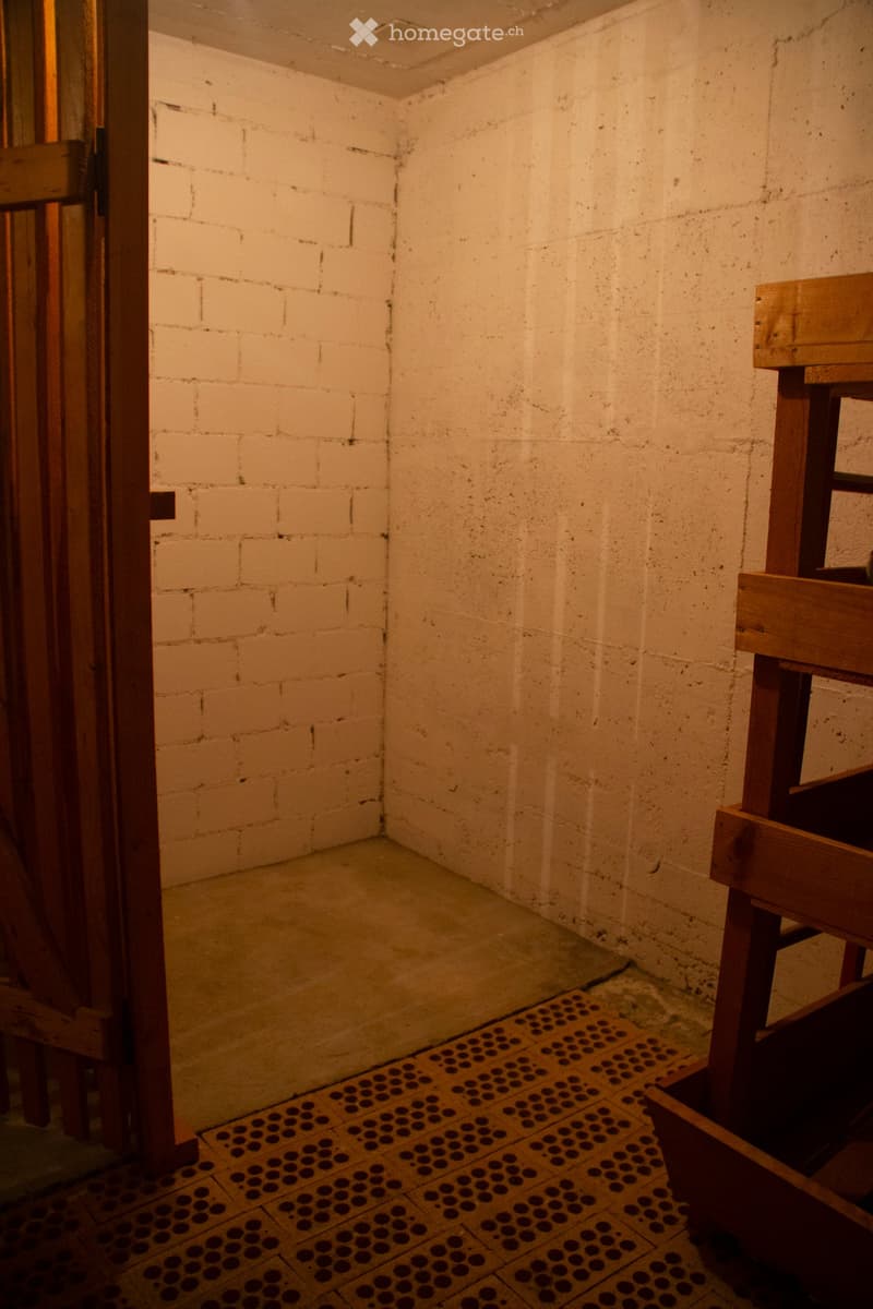 Empty room with exposed brick walls, concrete floor, and wooden door frame. A wooden stool is partially visible on the right side.