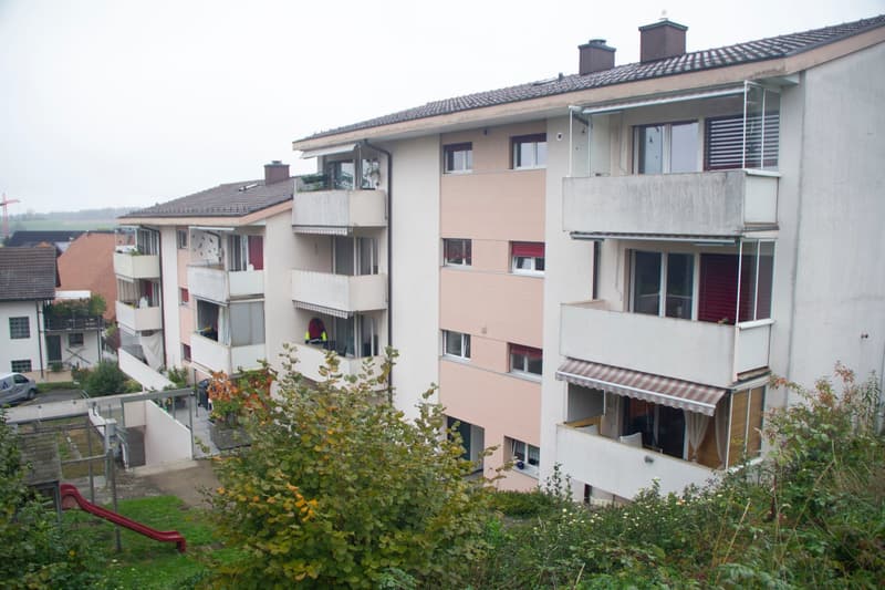 Multi-story residential building with balconies, windows, and awnings. The building has a light pink facade and is surrounded by greenery and a small playground area.