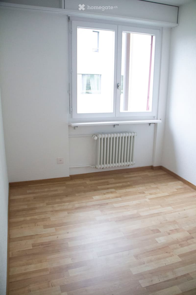Empty room with wooden flooring, white walls, a radiator, and a large window with a white frame and a shelf above it.