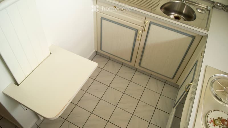 A small kitchen with white cabinets, a stainless steel sink, and a stove with a pot on it. The floor is tiled, and there is a white countertop visible.