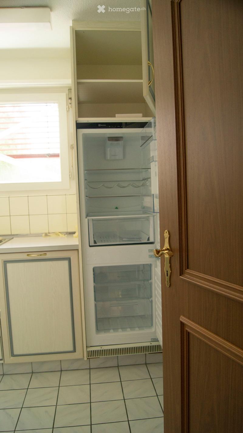 A small kitchen area with a white refrigerator and light-colored cabinets, featuring tiled flooring and a window with blinds.