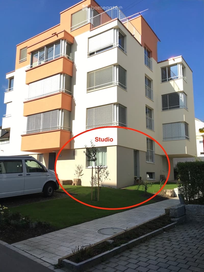 Modern two-story building with white and orange facade, multiple windows, balconies, and a small garden area in front.