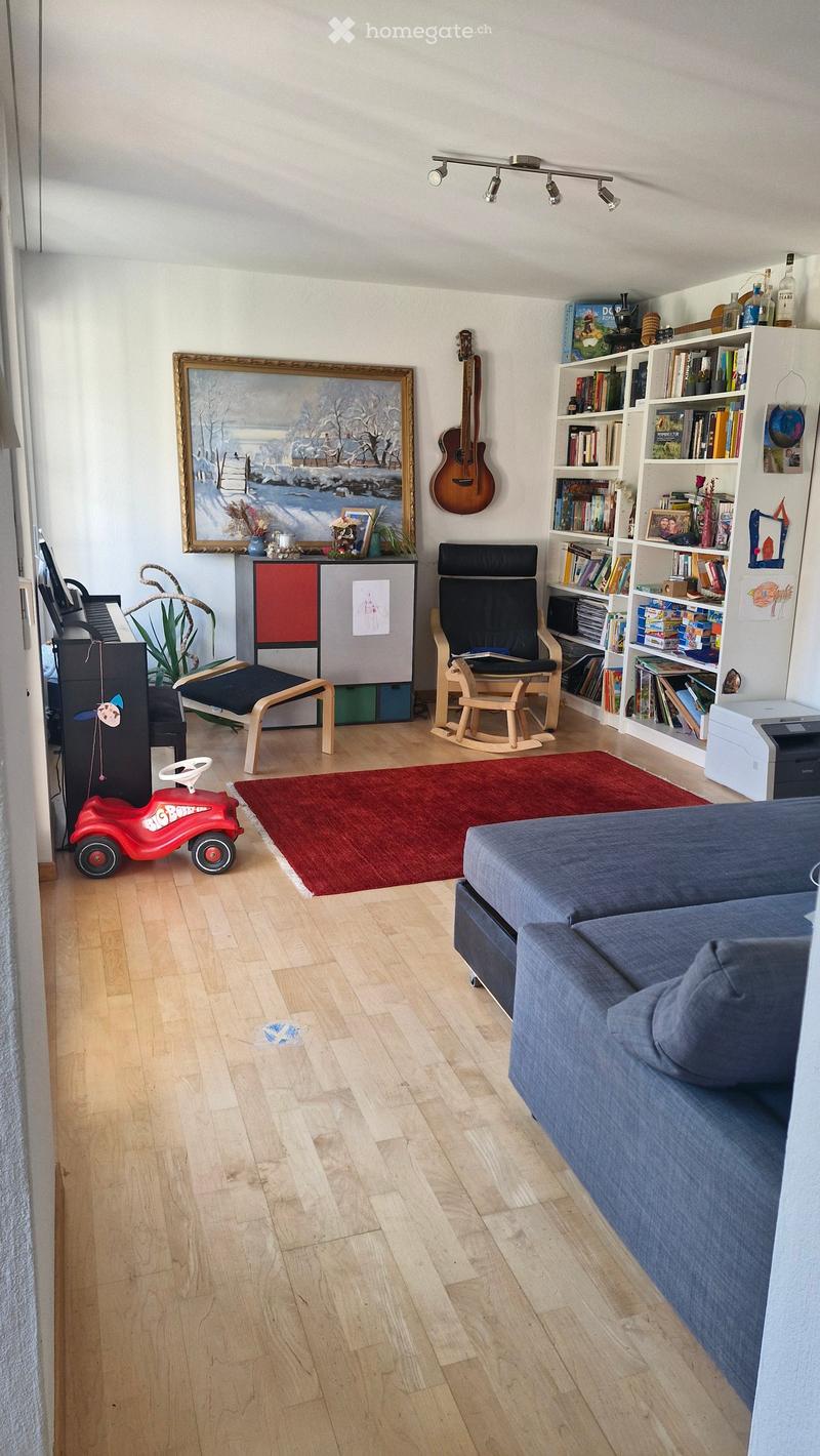 A cozy living room with a blue sofa, wooden rocking chair, bookshelf, guitar, and a red rug on the floor.