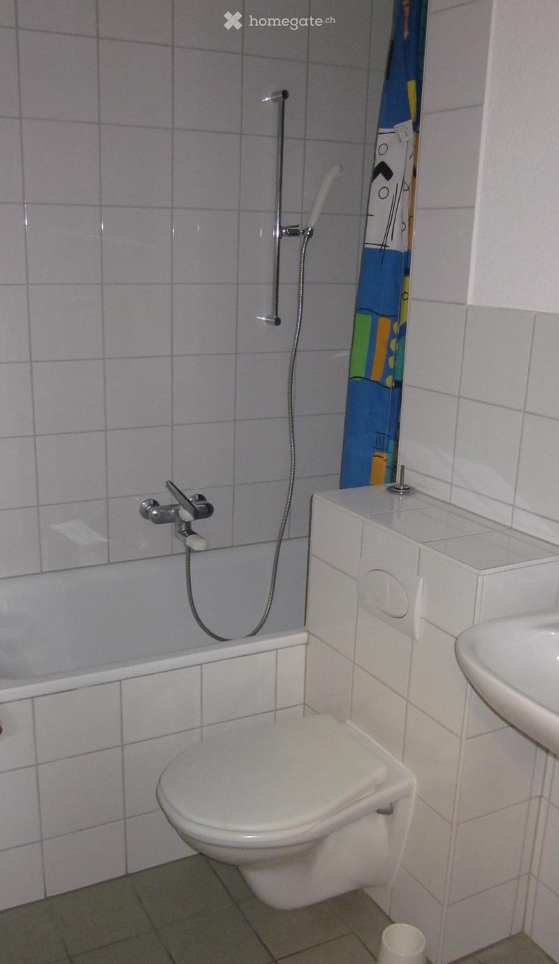 A small bathroom with a toilet, a sink, a faucet, and a cup on the floor. The walls are tiled, and there is a showerhead visible on the wall.