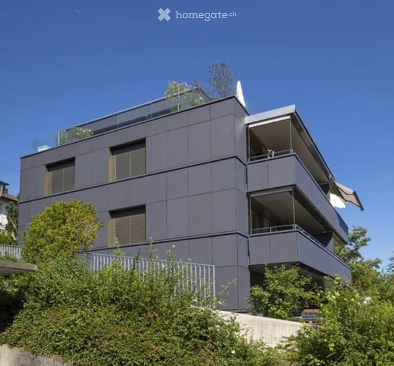 Modern multi-story building with dark grey facade, multiple balconies, and greenery around the base.