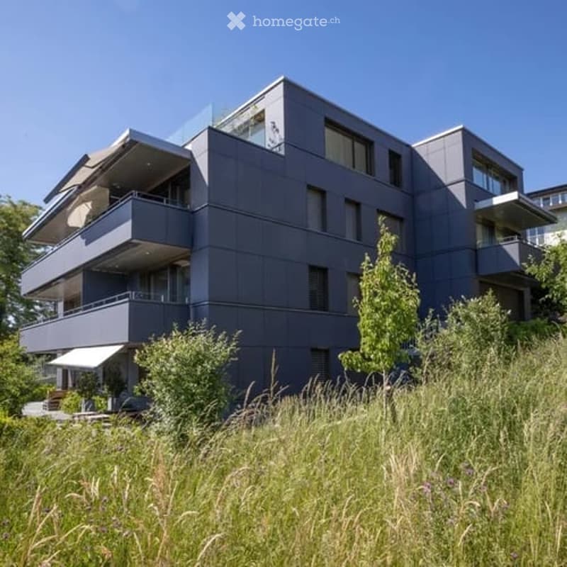 Modern multi-story building with grey facade, large windows, and multiple balconies overlooking a green landscape.
