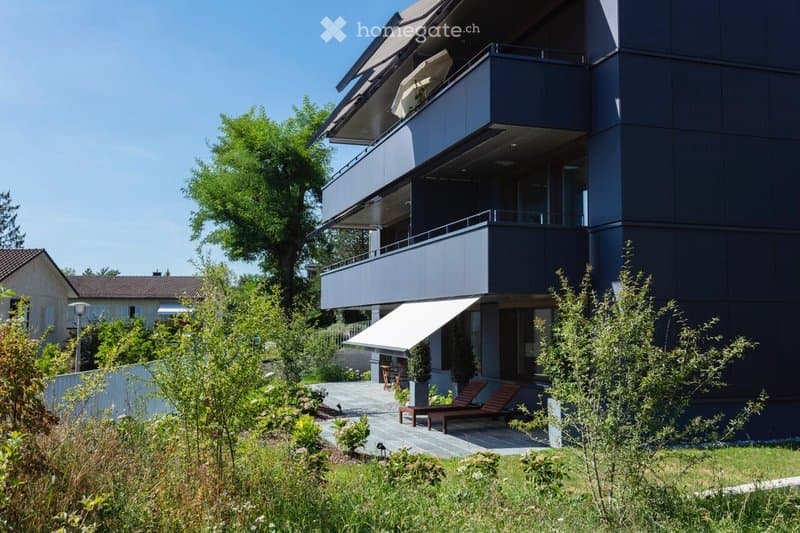 Modern multi-story building with balconies, surrounded by greenery and outdoor seating area.