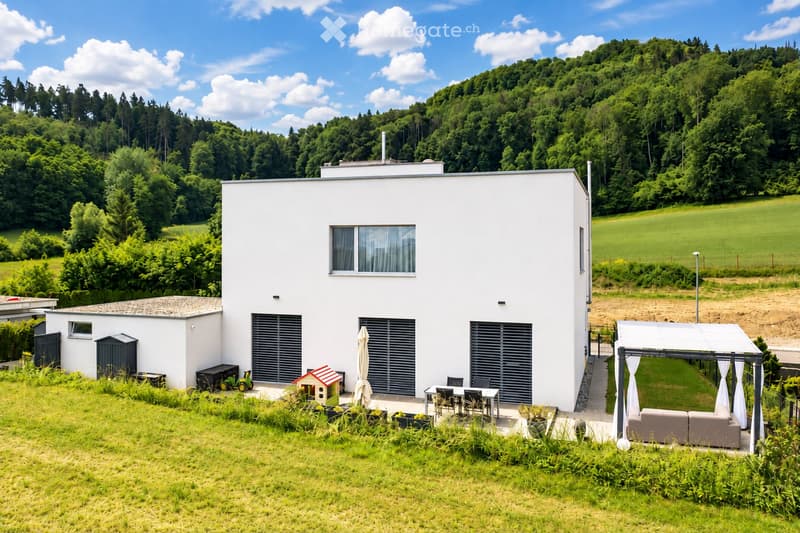Modern white house with large windows, garage, patio area with outdoor furniture, and scenic hillside backdrop.