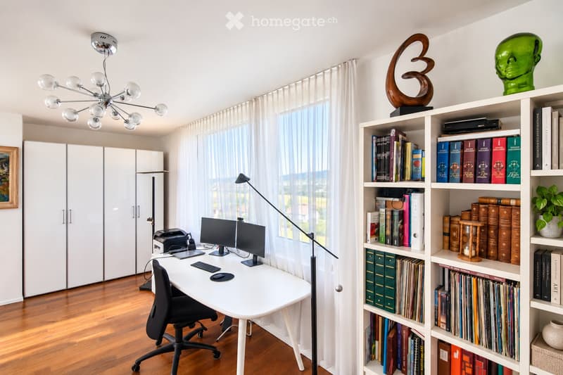 A modern, well-lit study or home office with a white desk, ergonomic chair, dual monitors, printer, and a large bookshelf filled with books and decorative items. The room features wooden flooring, large windows with sheer curtains, and a stylish chandelie