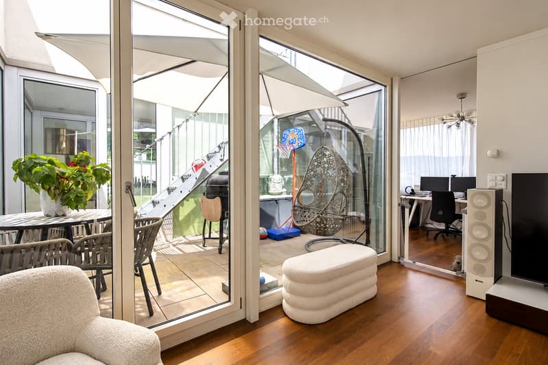 Modern living room with large glass doors opening to a balcony, featuring a white sofa, dining table with chairs, wicker hanging chair, basketball hoop, and a workspace with a laptop and speaker system.