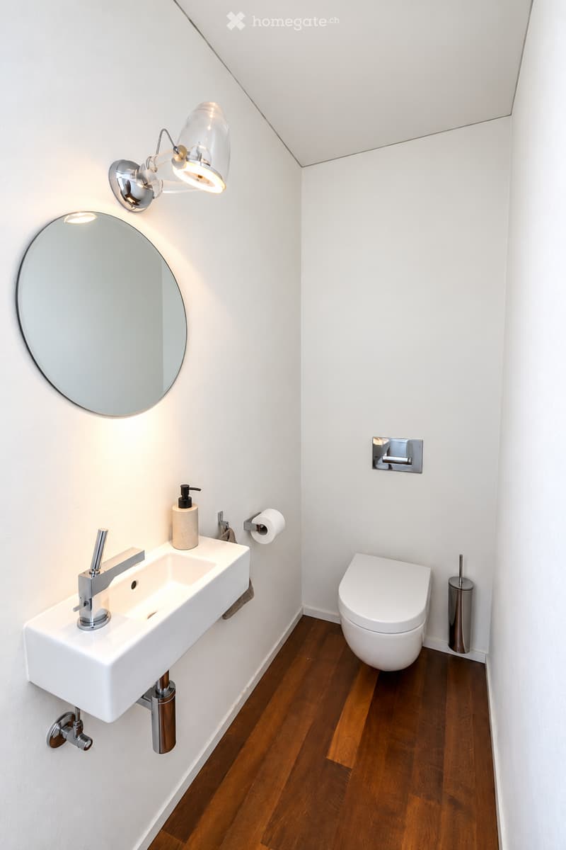 Modern bathroom with white walls, wooden floor, white sink with chrome faucet, round mirror, toilet, and wall-mounted light fixture.