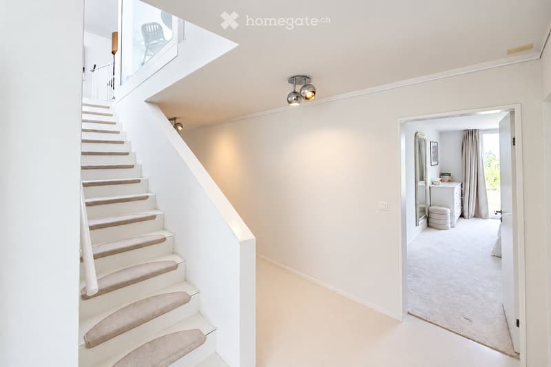 Modern hallway with a staircase leading to an upper level, featuring white walls, carpeted stairs, and a view into a bedroom with a vanity and mirror.
