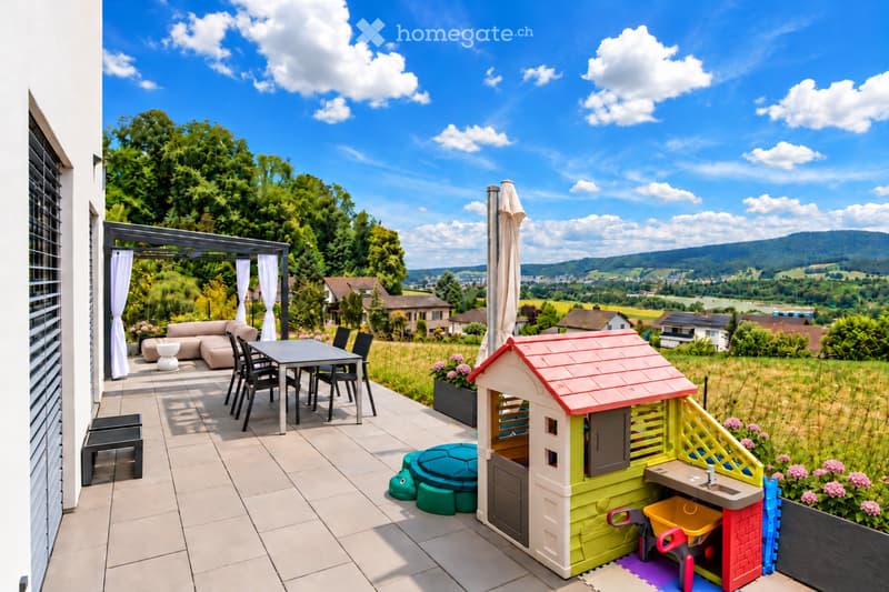 Balcony with a view of the countryside, featuring a sofa, table, chairs, a toy house, and a sink.