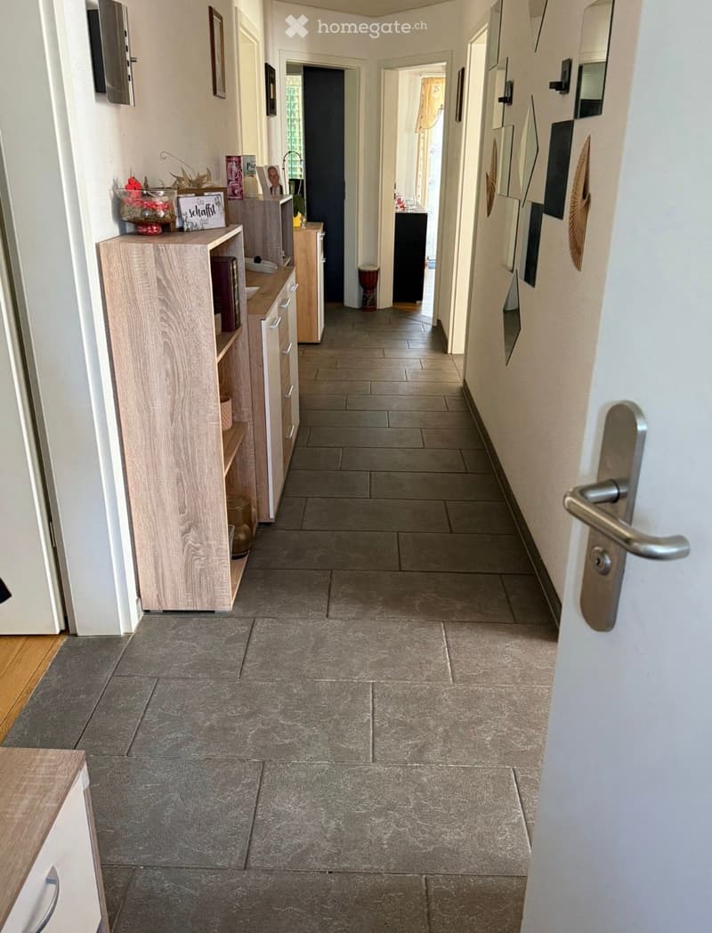 A clean, tiled hallway with light-colored walls, wooden cabinetry on the left, and several doors leading to other rooms.