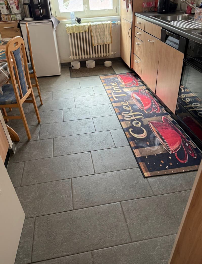Modern kitchen with tiled floor, wooden cabinets, stainless steel appliances, and a coffee-themed runner rug.