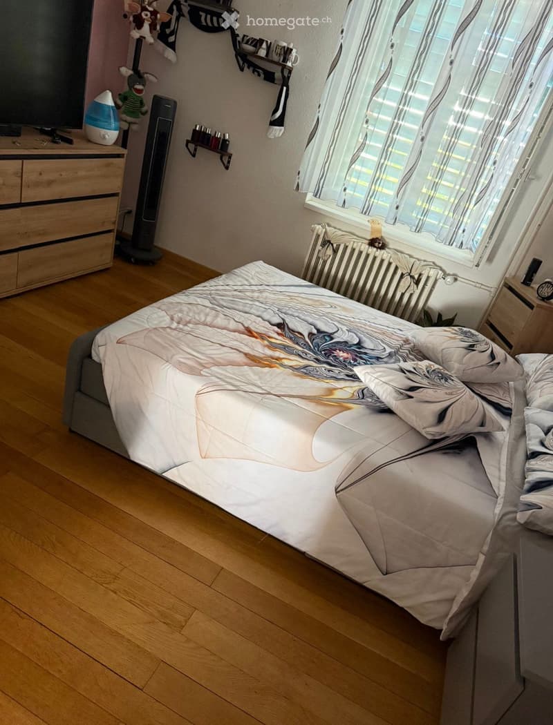 Cozy bedroom with wooden flooring, a large bed with a decorative duvet, nightstands, a fan, shelves with toys, and a window with curtains.
