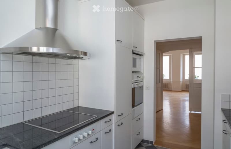 Modern kitchen with white cabinets, stainless steel appliances, and a black countertop. The room features a range hood, stove, oven, and microwave, with a view into an adjacent unfurnished room.