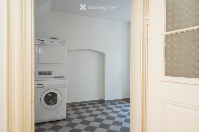 A small, well-lit room with a washing machine and dryer stacked vertically, featuring a checkered black and white tile floor and light blue walls.