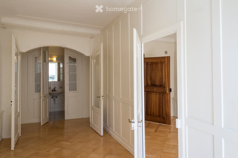 Elegant hallway with wooden flooring, white walls, and arched doorways leading to a bathroom and another room. The bathroom features a sink, mirror, and tiled walls.