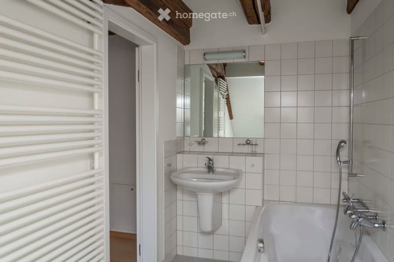 A modern bathroom with white tiled walls, a sink, a bathtub, a mirror, and a light fixture above the mirror. The room features exposed wooden beams on the ceiling and a white radiator on the left side.