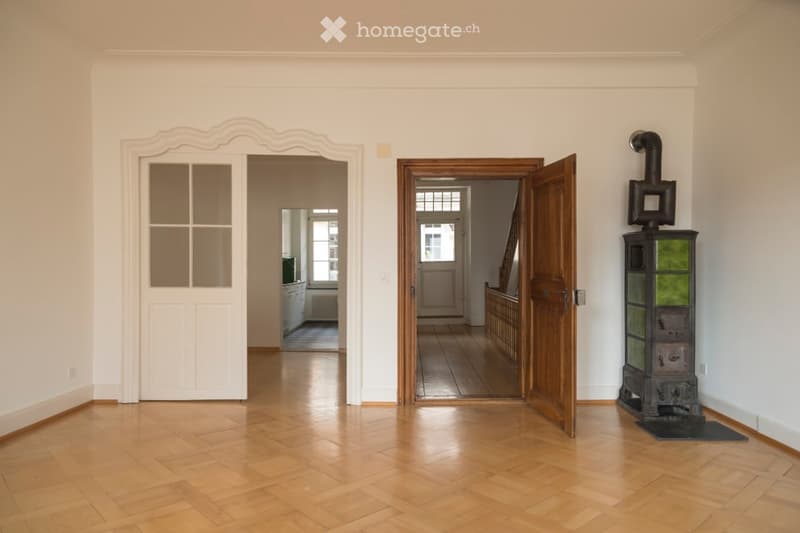 Bright hallway with wooden flooring, white walls, and two open doors leading to adjacent rooms. A vintage stove is visible on the right side.