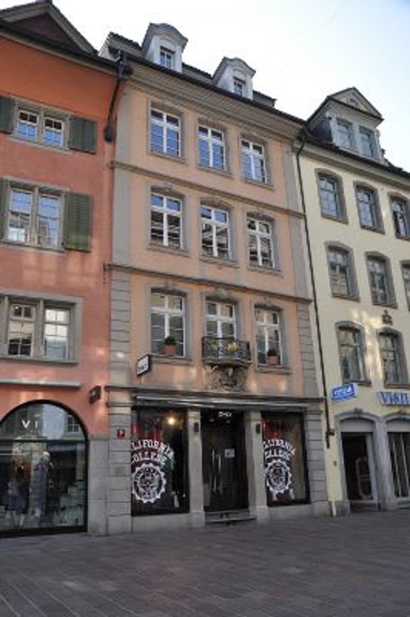 Three-story urban building with classic European architecture, featuring large windows, decorative balconies, and storefronts at ground level.