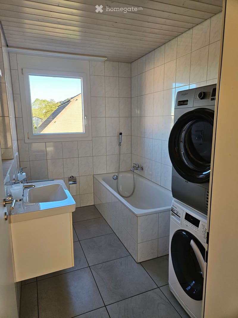 Modern bathroom with a white bathtub, sink, washing machine, and a window offering a view of the roof and surrounding greenery.