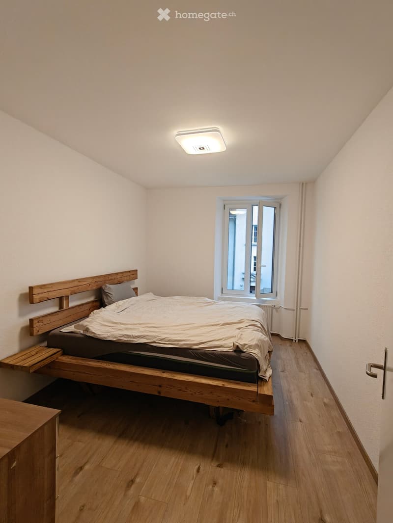 Cozy bedroom with wooden bed, headboard, and pillow, featuring wooden flooring and a window with white frame.