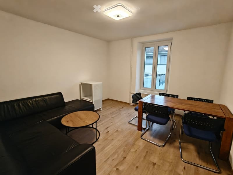 A modern, well-lit room with a black leather sofa, a small round wooden coffee table, a wooden dining table with four chairs, and a white shelving unit.