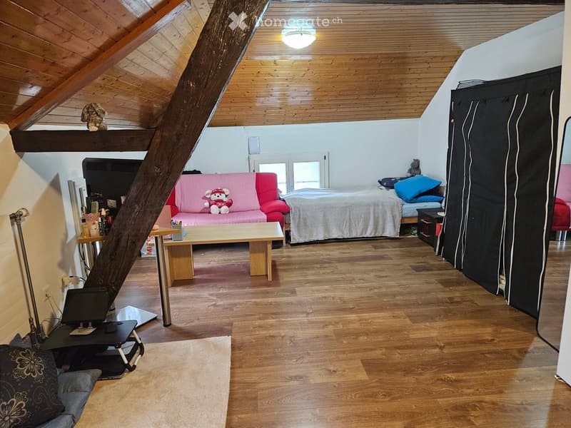 A cozy attic room with wooden floors and slanted wooden ceilings. Features include a pink sofa with a teddy bear, a wooden coffee table, a bed with pillows and a blanket, a laptop on a small table, and a large window letting in natural light.