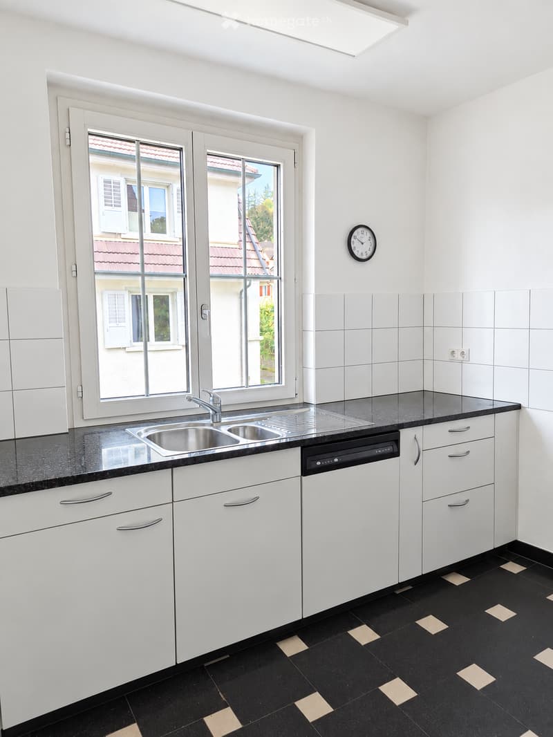 Modern kitchen with white cabinets, black countertops, stainless steel sink, and a dishwasher. Features a large window with a view of a neighboring building.