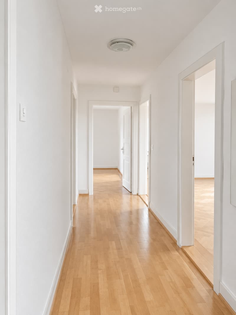 A long, narrow hallway with light wooden flooring and white walls, featuring several open doors leading to other rooms.