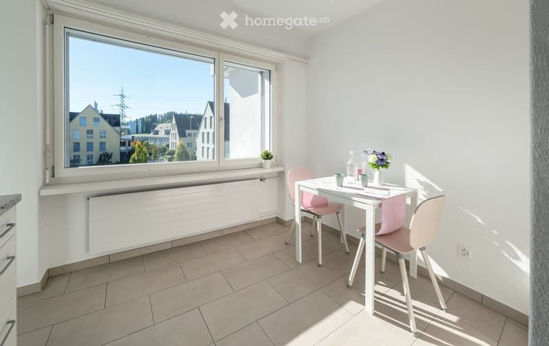 Bright room with tiled floor, white walls, large windows, a small table with chairs, and a vase with flowers on the table.