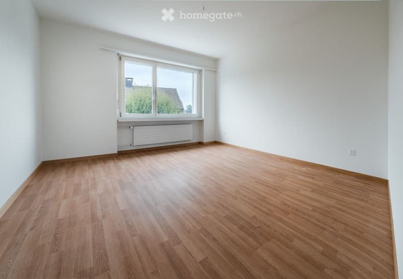 Empty room with wooden flooring, white walls, and a large window with white frames and blinds. A radiator is visible below the window.