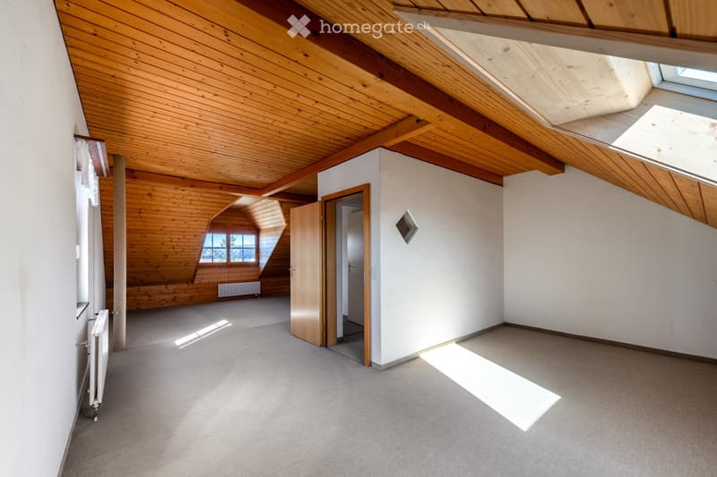 Empty attic space with wooden beams, skylights, and large windows, featuring a carpeted floor and white walls.