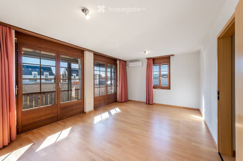 Empty room with wooden flooring, large wooden-framed windows, and pink curtains. Natural light streams in, illuminating the space.