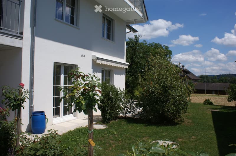 A well-maintained garden area with a white house in the background, featuring a variety of plants, a blue barrel, and a clear view of the surrounding landscape.