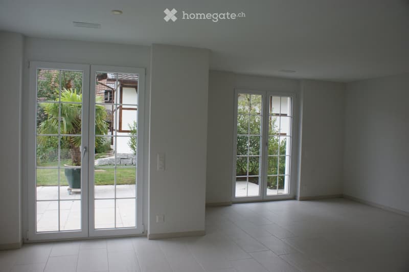 Empty room with large sliding glass doors leading to a garden, tiled floor, and neutral-colored walls.
