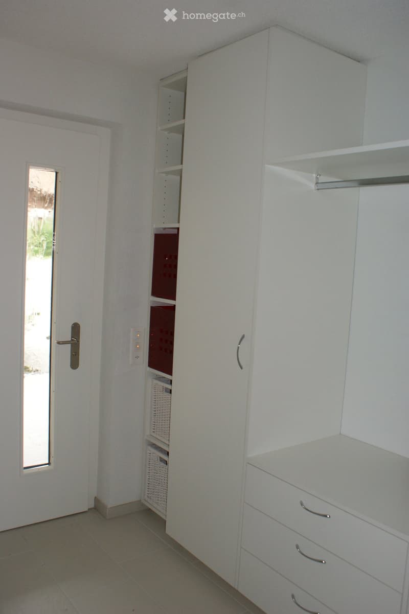 Empty room with white walls, tiled floor, built-in white wardrobe with shelves and drawers, and a small windowed door leading outside.