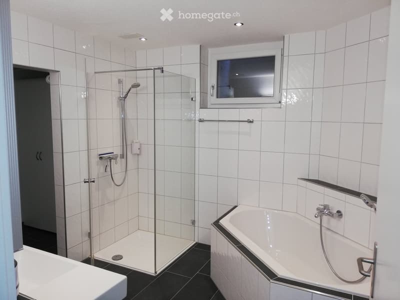 Modern bathroom with white tiles, a bathtub, a glass-enclosed shower, and a window above the bathtub.
