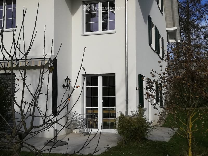 White house exterior with a small patio area featuring a table and chairs, surrounded by bare trees and greenery.
